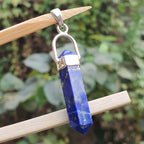 Blue crystal pendant with silver hook on a wooden stick against a green leafy background