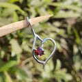 Heart-shaped silver pendant with a red gemstone on a stick against a blurred natural background