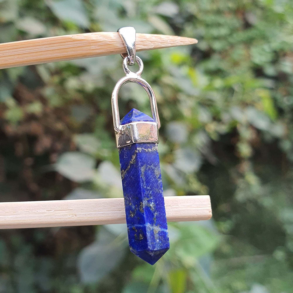 Blue crystal pendant with silver chain on a wooden stick against a blurred green background