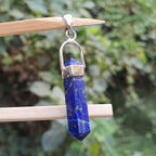 Blue crystal pendant with silver chain on a wooden stick against a blurred green background
