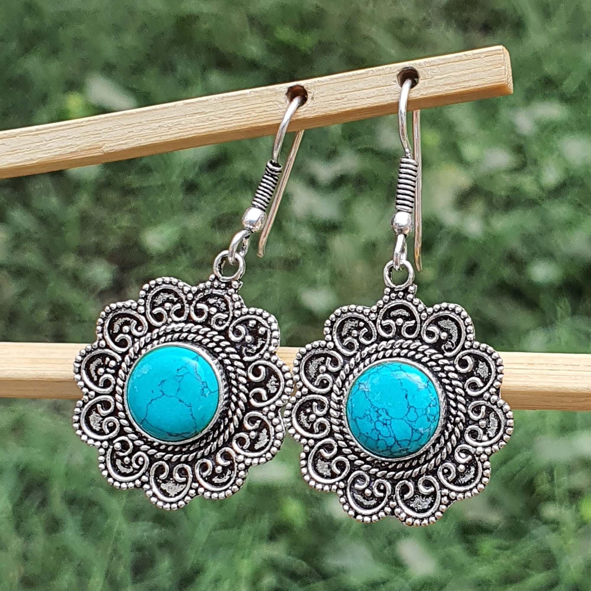 Pair of earrings with turquoise stones and silver filigree on a wooden stick against a blurred green background.