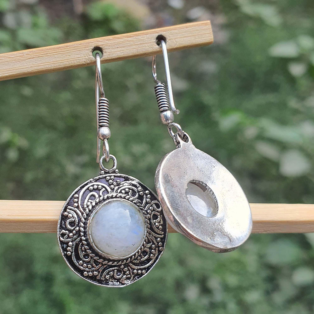 Silver earrings with circular designs and a central gemstone on a wooden stick against a blurred green background.
