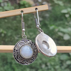 Silver earrings with circular designs and a central gemstone on a wooden stick against a blurred green background.