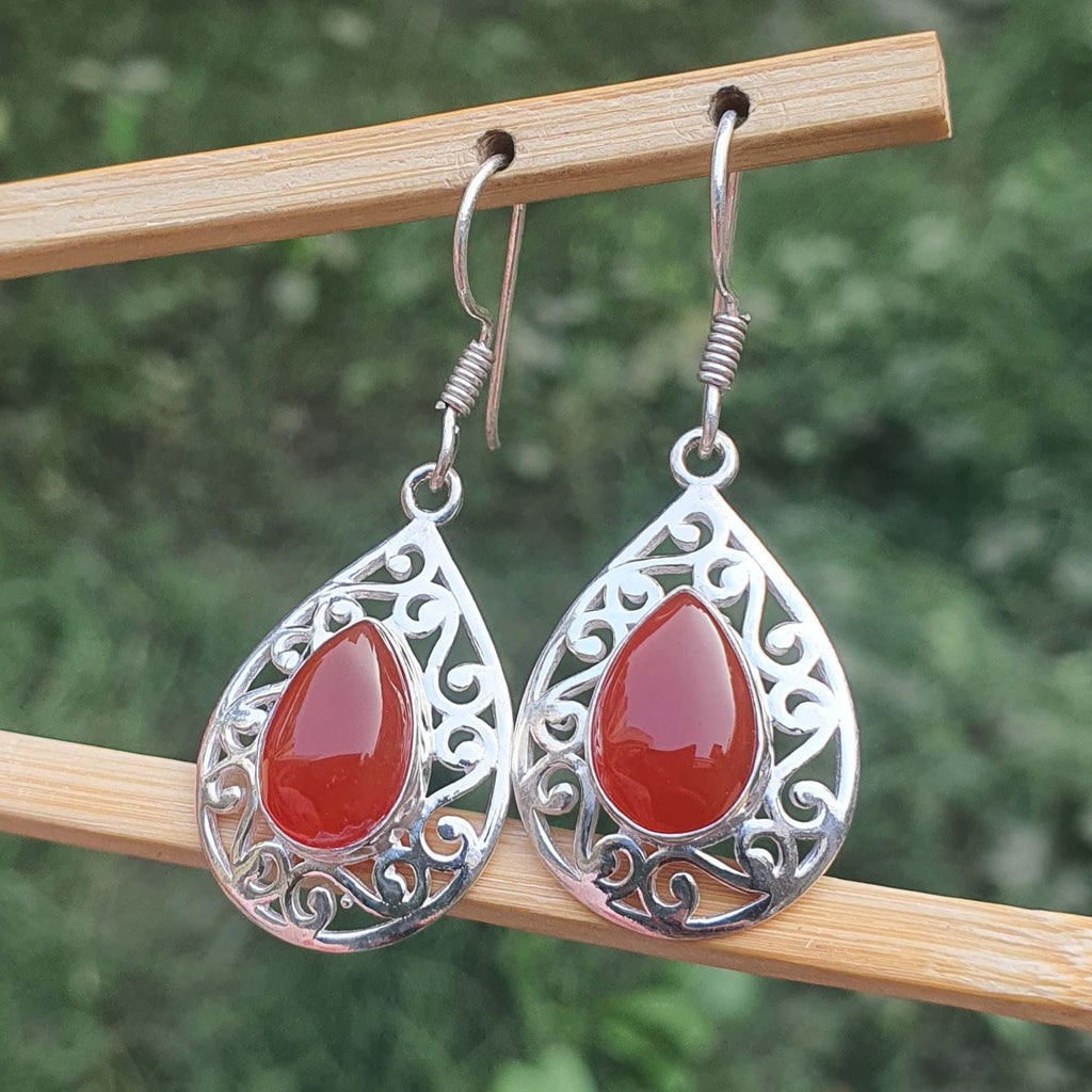 Teardrop-shaped earrings with red stones and silver filigree on a wooden stick against a blurred green background