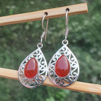 Teardrop-shaped earrings with red stones and silver filigree on a wooden stick against a blurred green background