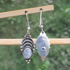 Two silver earrings with gemstones hanging on a wooden stick against a blurred green background.