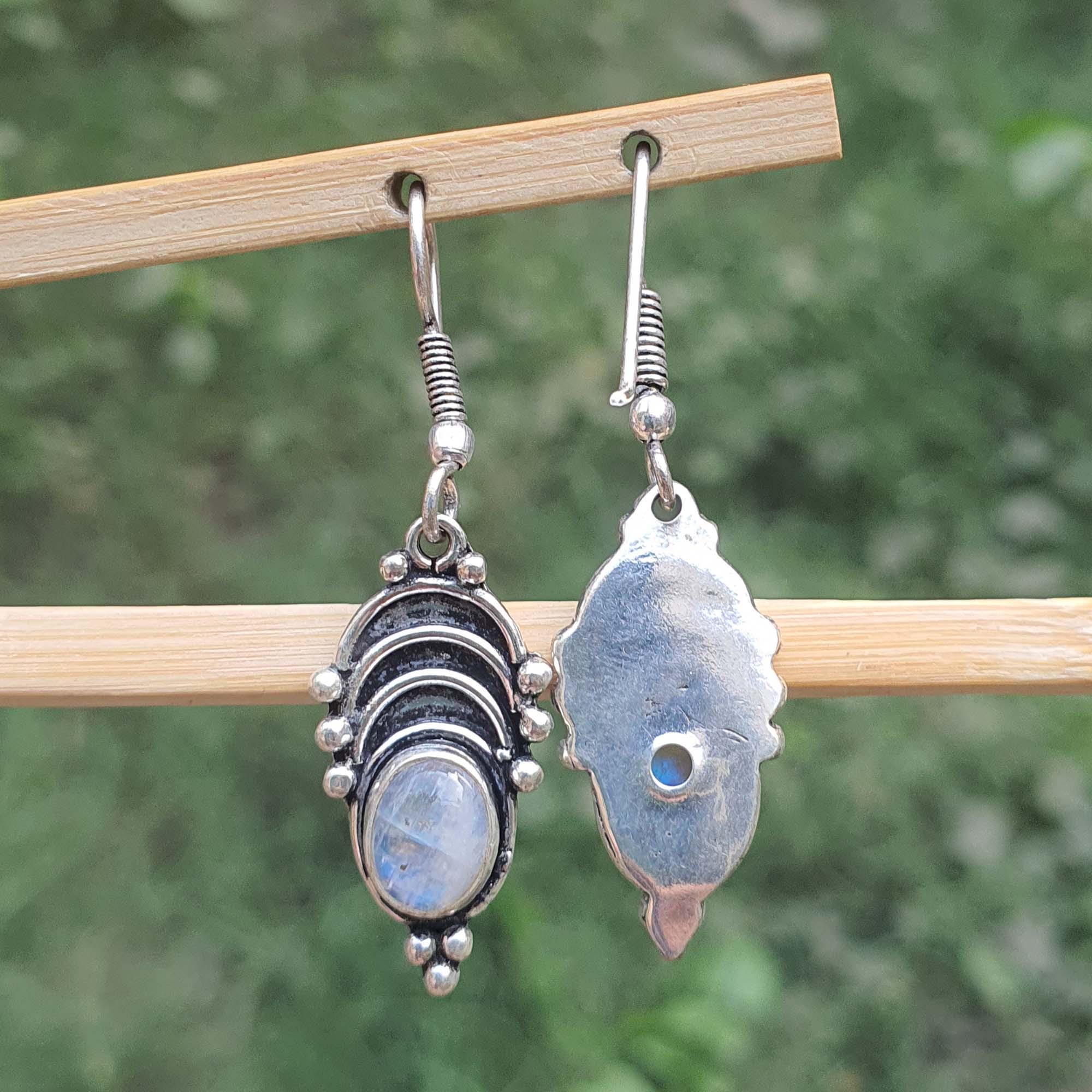 Two silver earrings with gemstones hanging on a wooden stick against a blurred green background.