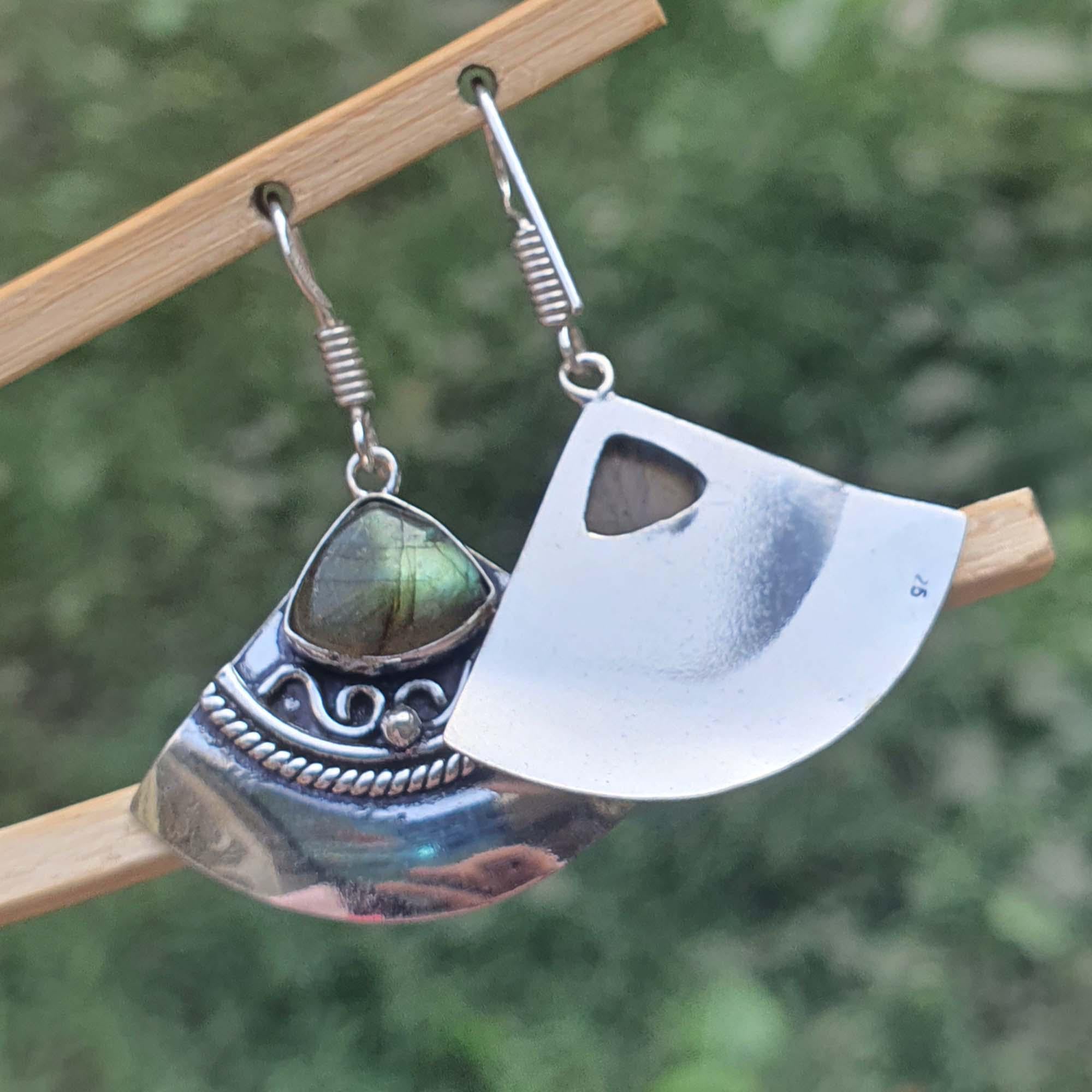 Silver axe-shaped earrings with a gemstone on a wooden stand against a blurred green background