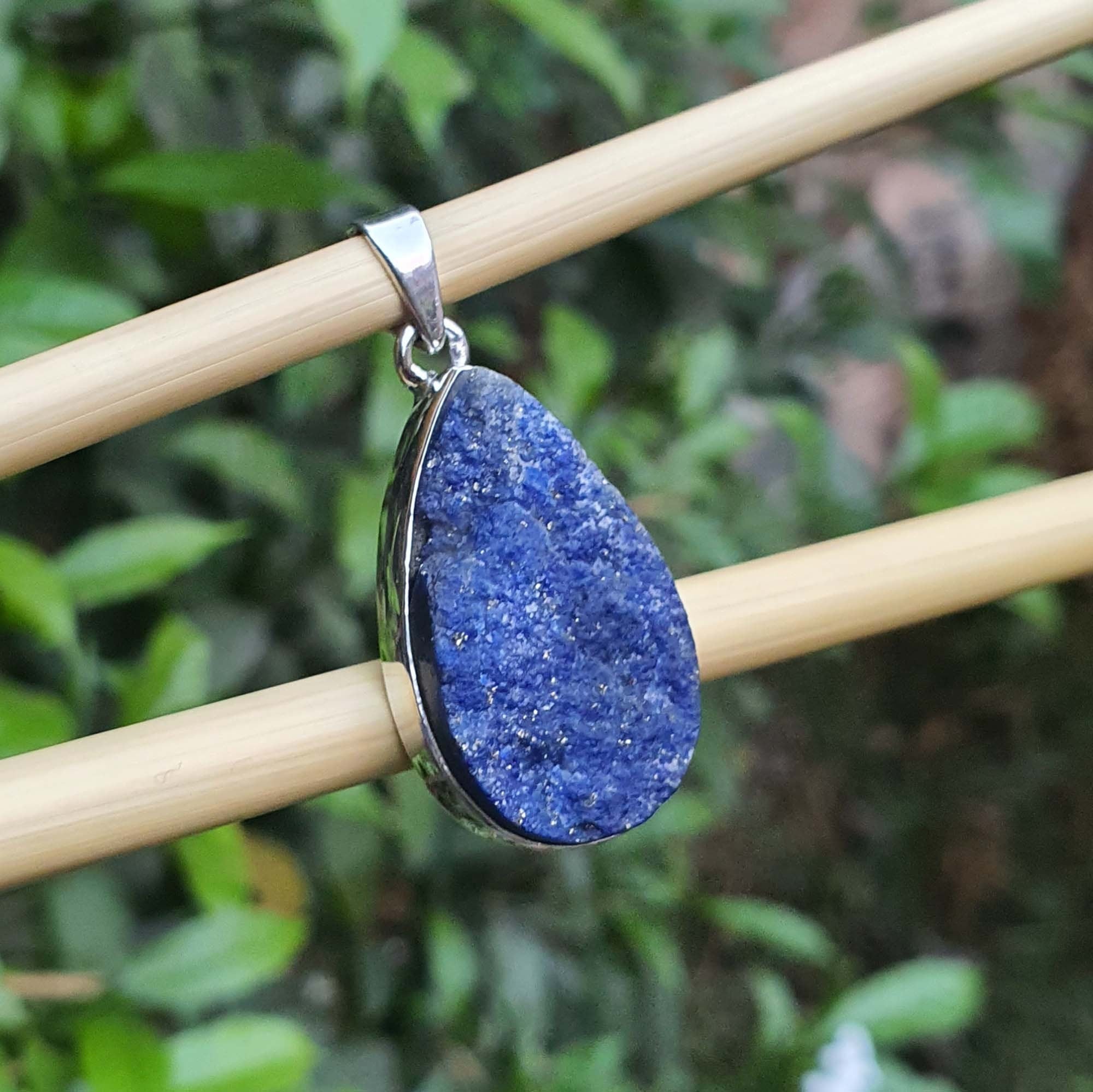 Blue pendant with silver frame on a wooden stick against a green leafy background