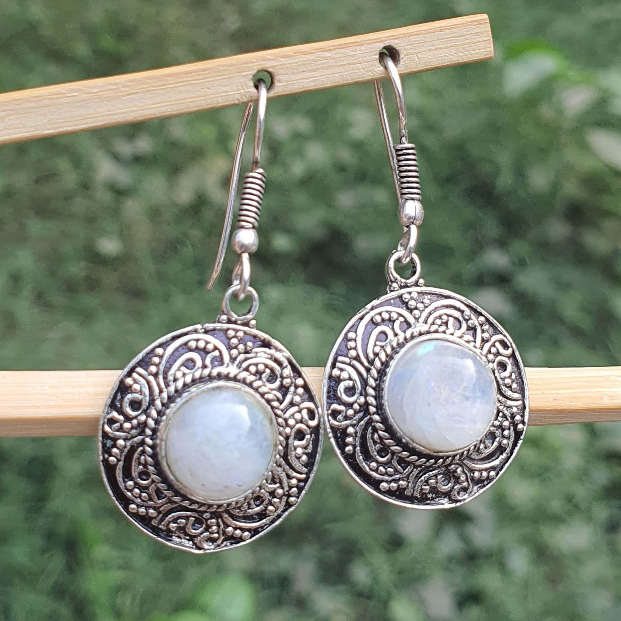 Silver earrings with circular designs and light-colored stones on a wooden stand against a blurred green background.