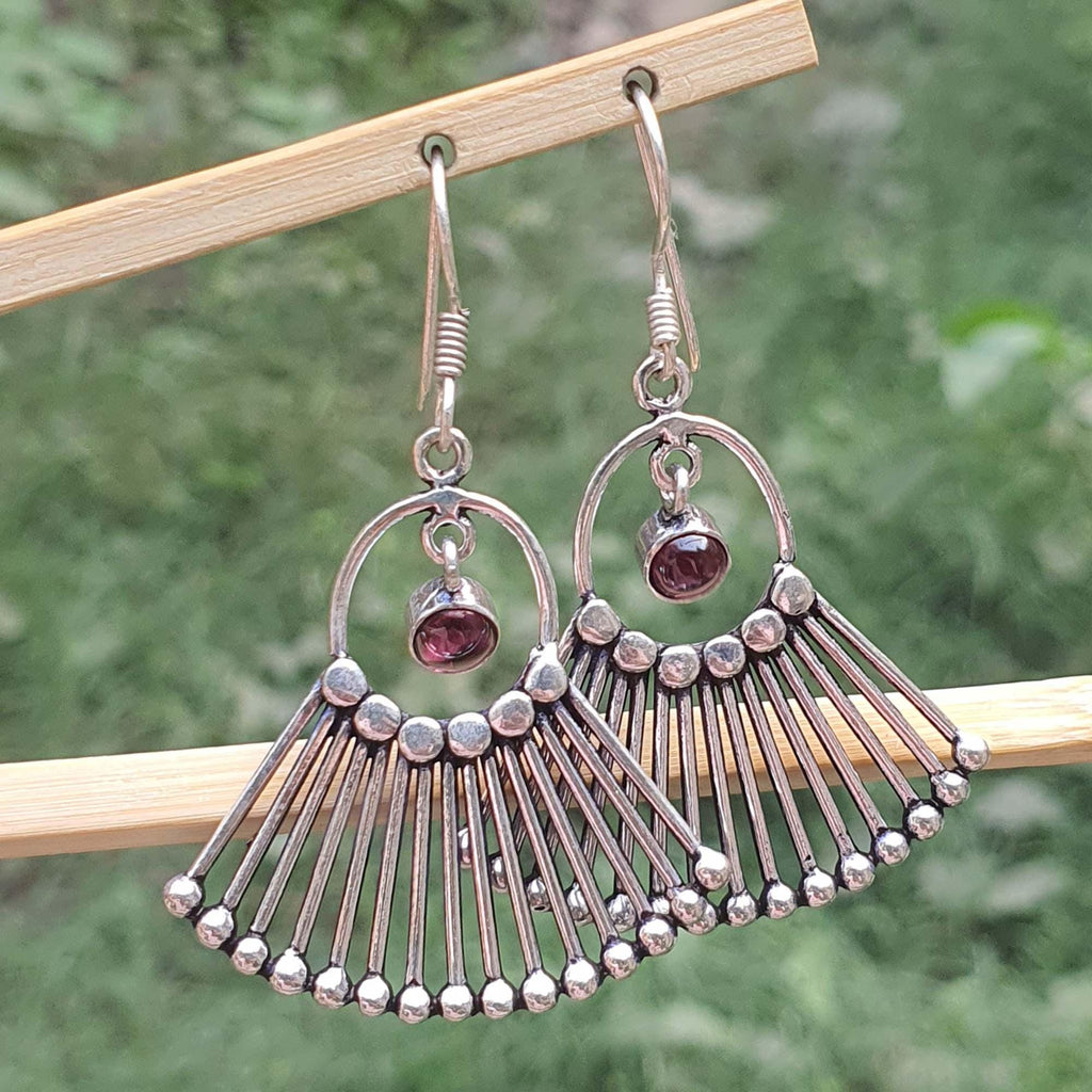 Silver earrings with fan-like design and red gemstones hanging on a wooden stick against a blurred green background.