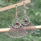 Silver earrings with fan-like design and red gemstones hanging on a wooden stick against a blurred green background.