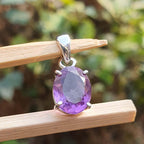 Purple gemstone pendant on a silver chain held by wooden sticks with a blurred green background