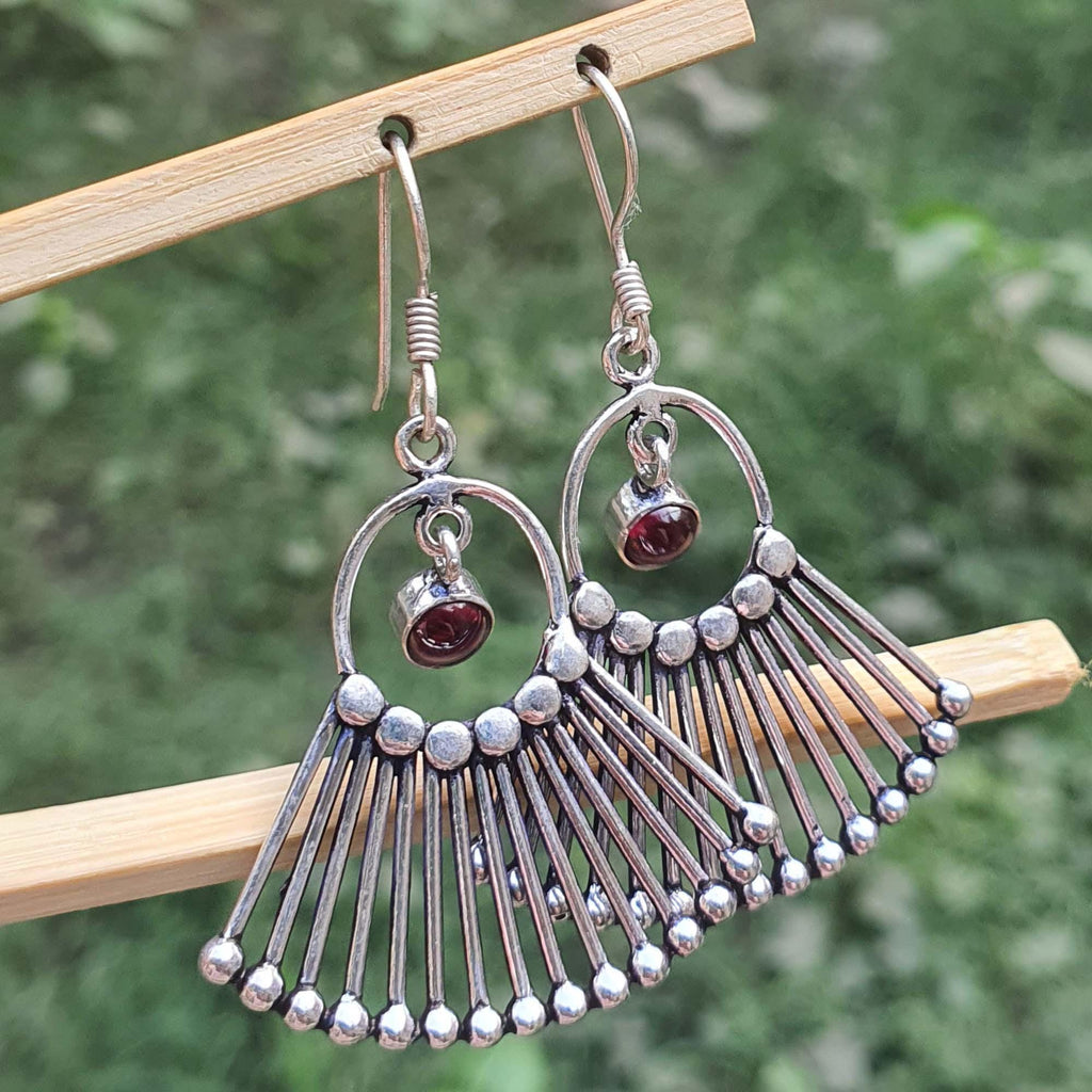 Silver earrings with fan-like design and red gemstones hanging on a wooden stick against a blurred green background.
