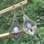 Ornate silver earrings with gemstones on a wooden stick against a green background