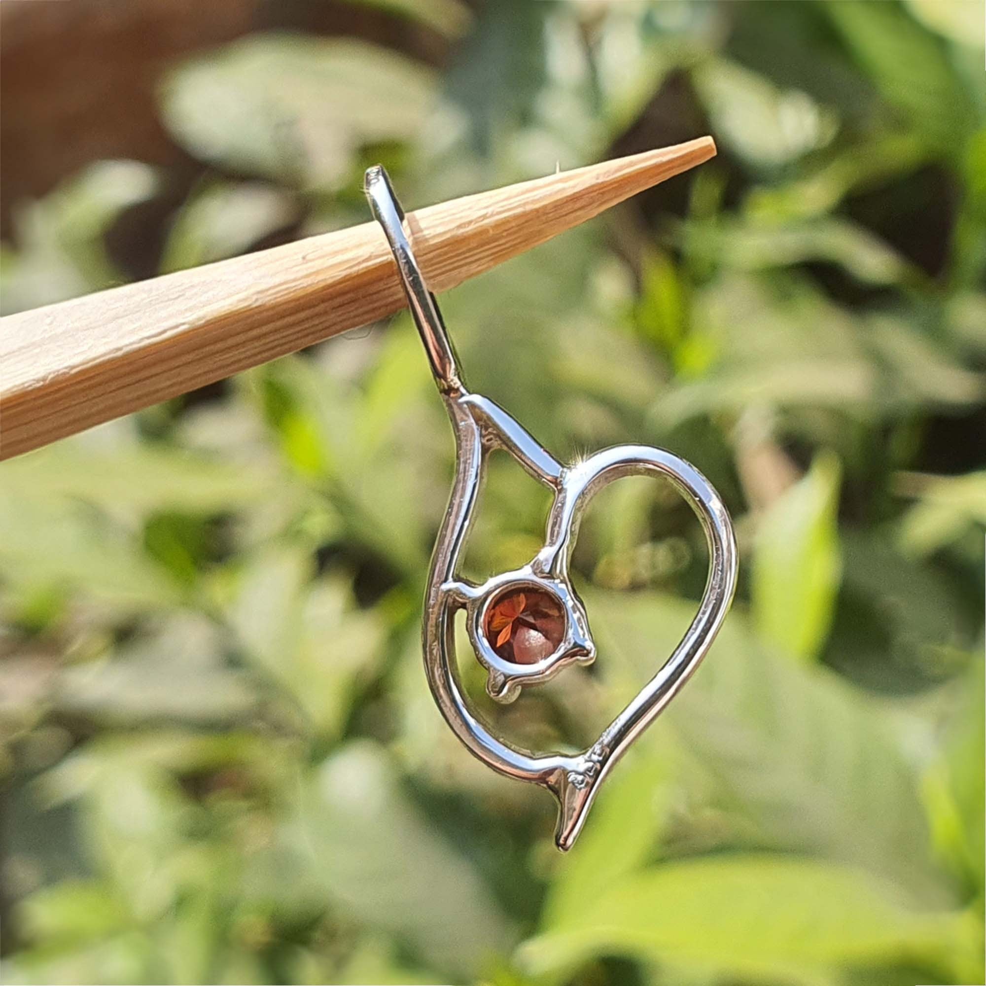 Silver pendant with a heart design and embedded gemstone on a wooden stick against a blurred green background