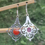 Ornate silver earrings with red and purple stones on a wooden stand against a blurred green background