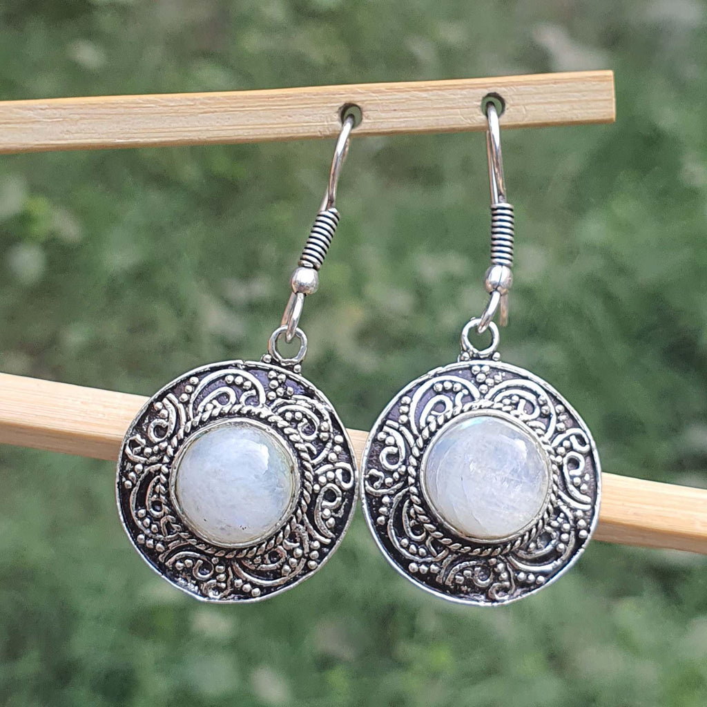 Silver earrings with circular designs and white stones on a wooden stick against a blurred green background