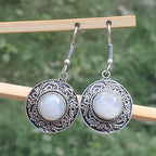 Silver earrings with circular designs and white stones on a wooden stick against a blurred green background