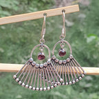 Silver earrings with fan-like design and red gemstones on a wooden stick against a green background