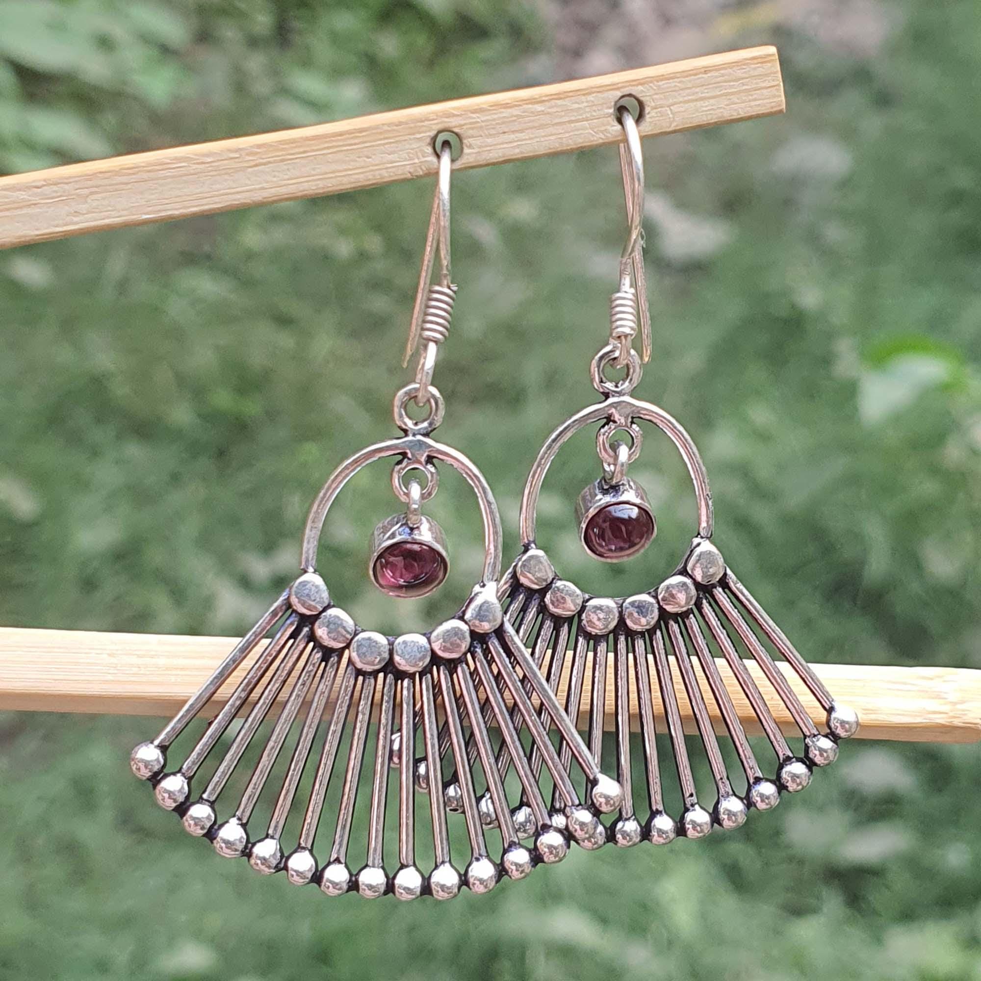 Silver earrings with fan-like design and red gemstones on a wooden stick against a green background