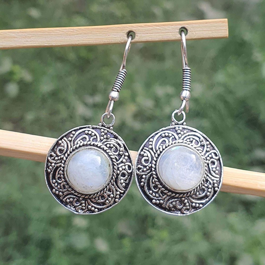 Silver earrings with circular designs and light-colored stones on a wooden stick against a blurred green background.