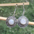Silver earrings with circular designs and light-colored stones on a wooden stick against a blurred green background.