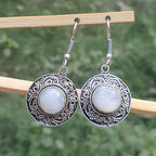 Silver earrings with circular designs and light-colored stones on a wooden stick against a blurred green background.