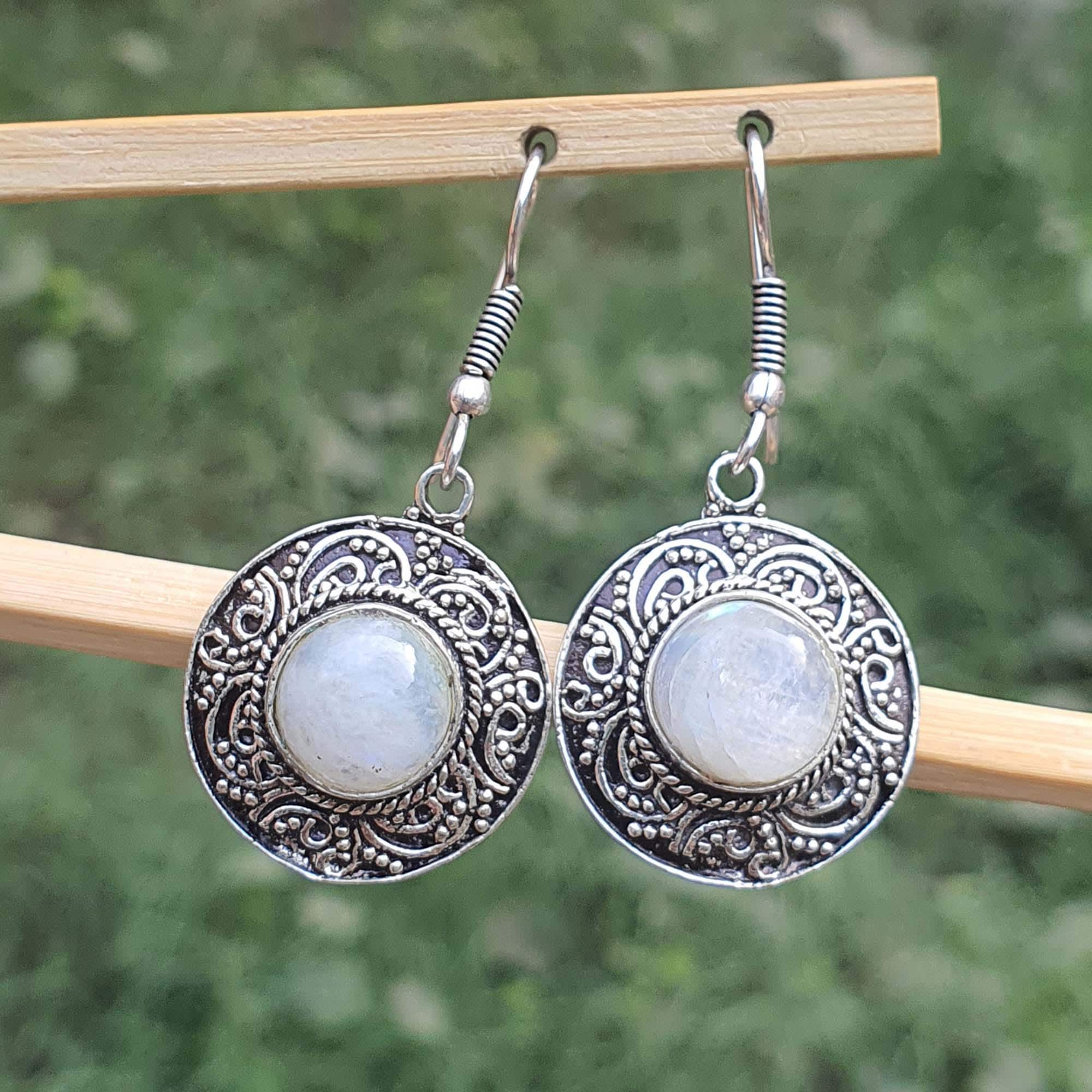 Silver earrings with circular designs and light-colored stones on a wooden stick against a blurred green background.