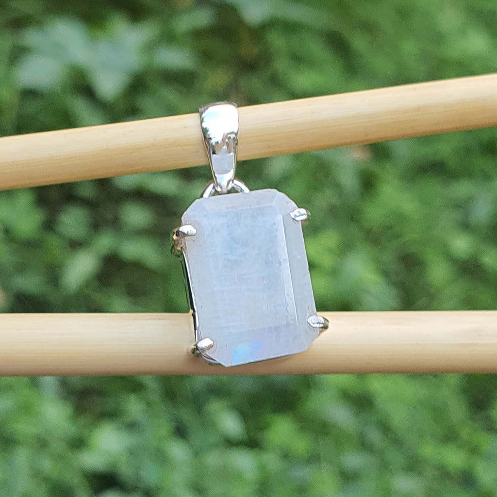 Silver pendant with a rectangular gemstone on a wooden stick against a green foliage background