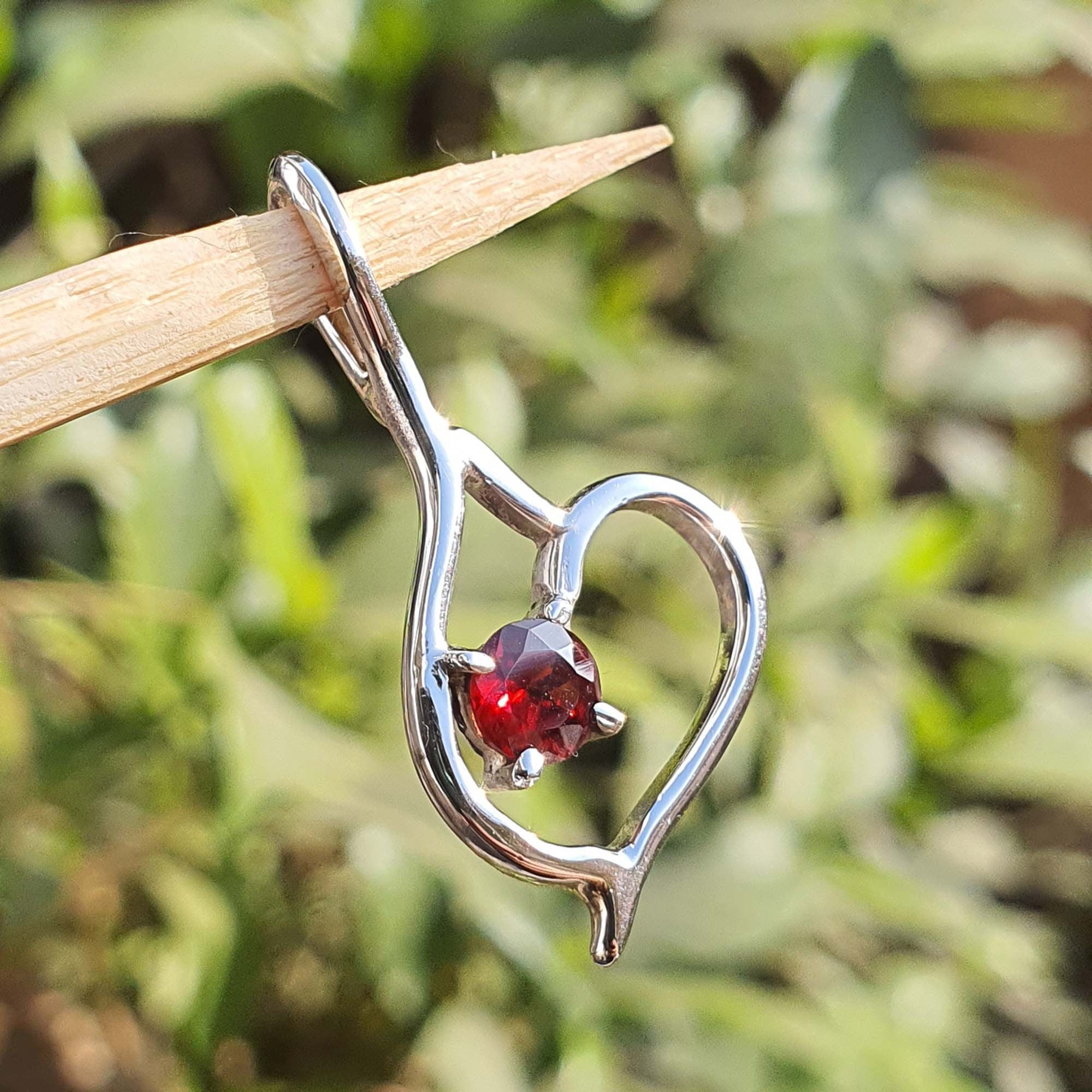 Silver pendant with a red gemstone held by a wooden stick against a blurred green background