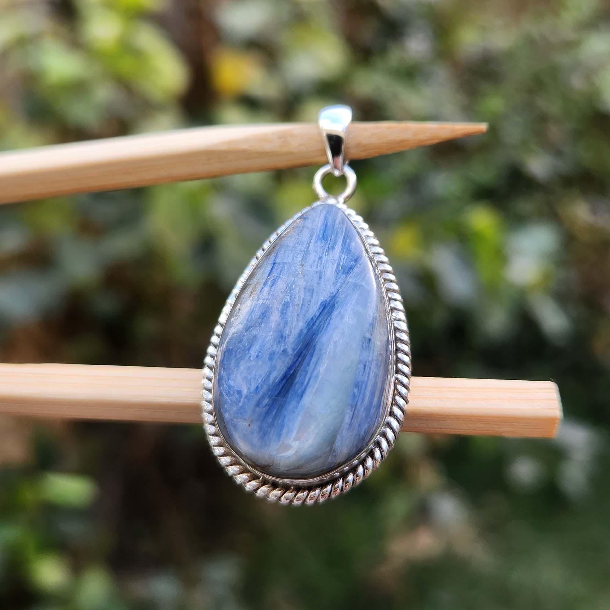 Blue teardrop pendant with silver frame on a wooden stick against a blurred green background