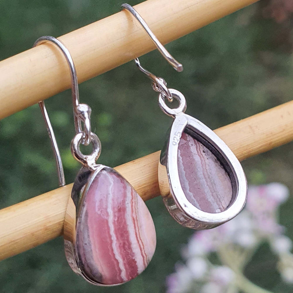 Teardrop-shaped pink stone earrings with silver hooks on a wooden stick against a blurred natural background.