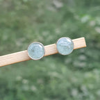 Pair of round earrings with a light blue gemstone on a wooden stick against a blurred green background