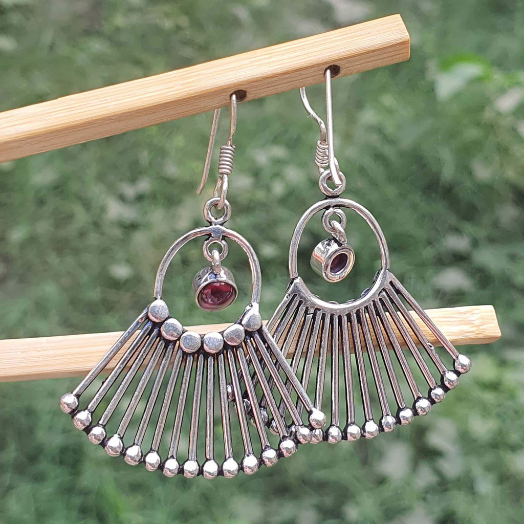Silver fan-shaped earrings with a central gemstone on a wooden stick against a blurred green background