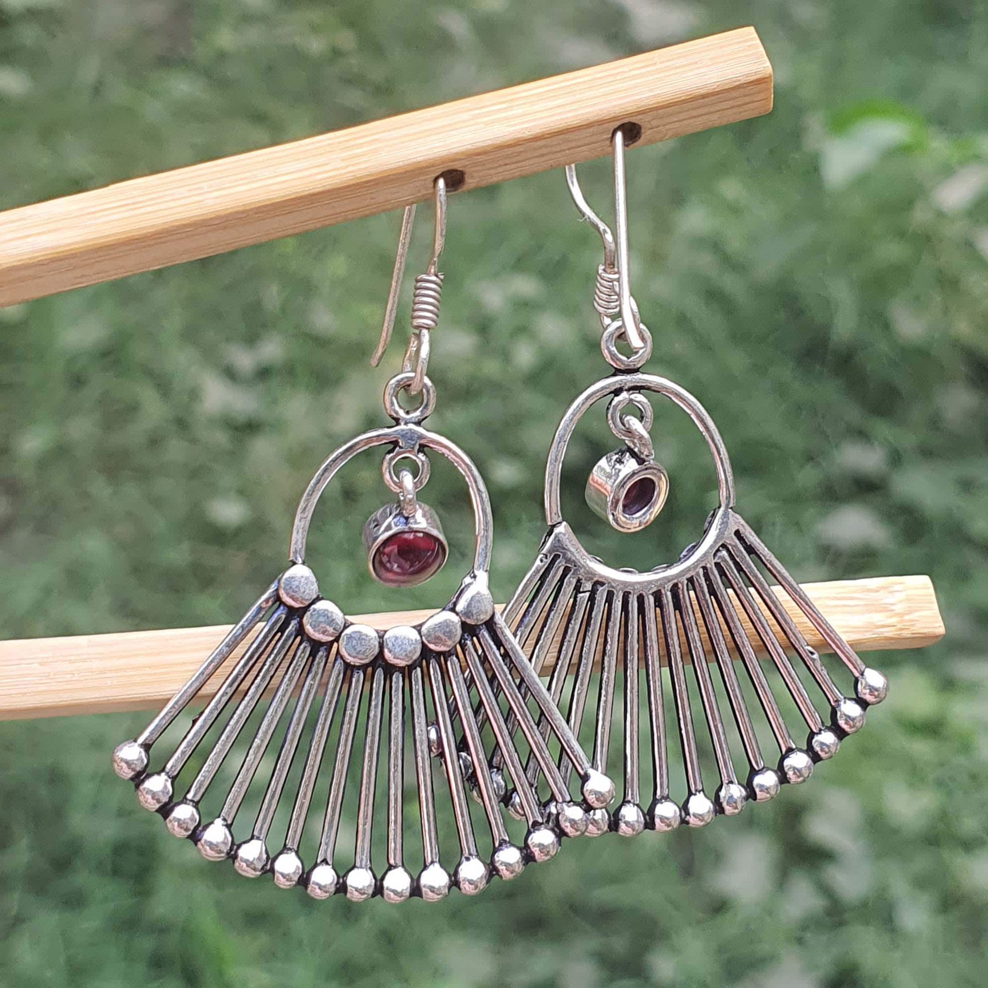 Silver fan-shaped earrings with a central gemstone on a wooden stick against a blurred green background