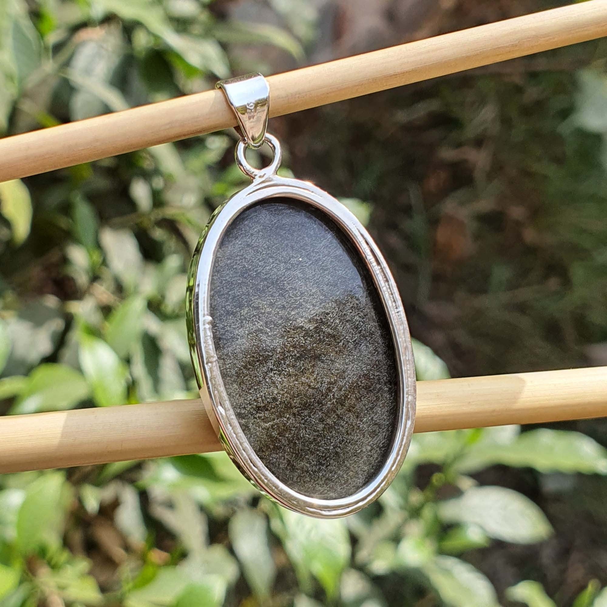 Pendant with a dark stone and silver frame on a wooden stick against a blurred green leafy background