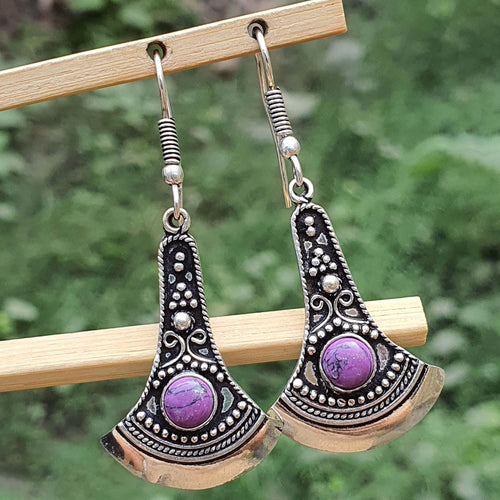 Pair of silver and purple earrings hanging on a wooden stick with a green blurred background.