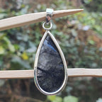 Teardrop-shaped pendant with dark stone and silver frame on a wooden stick against a blurred natural background