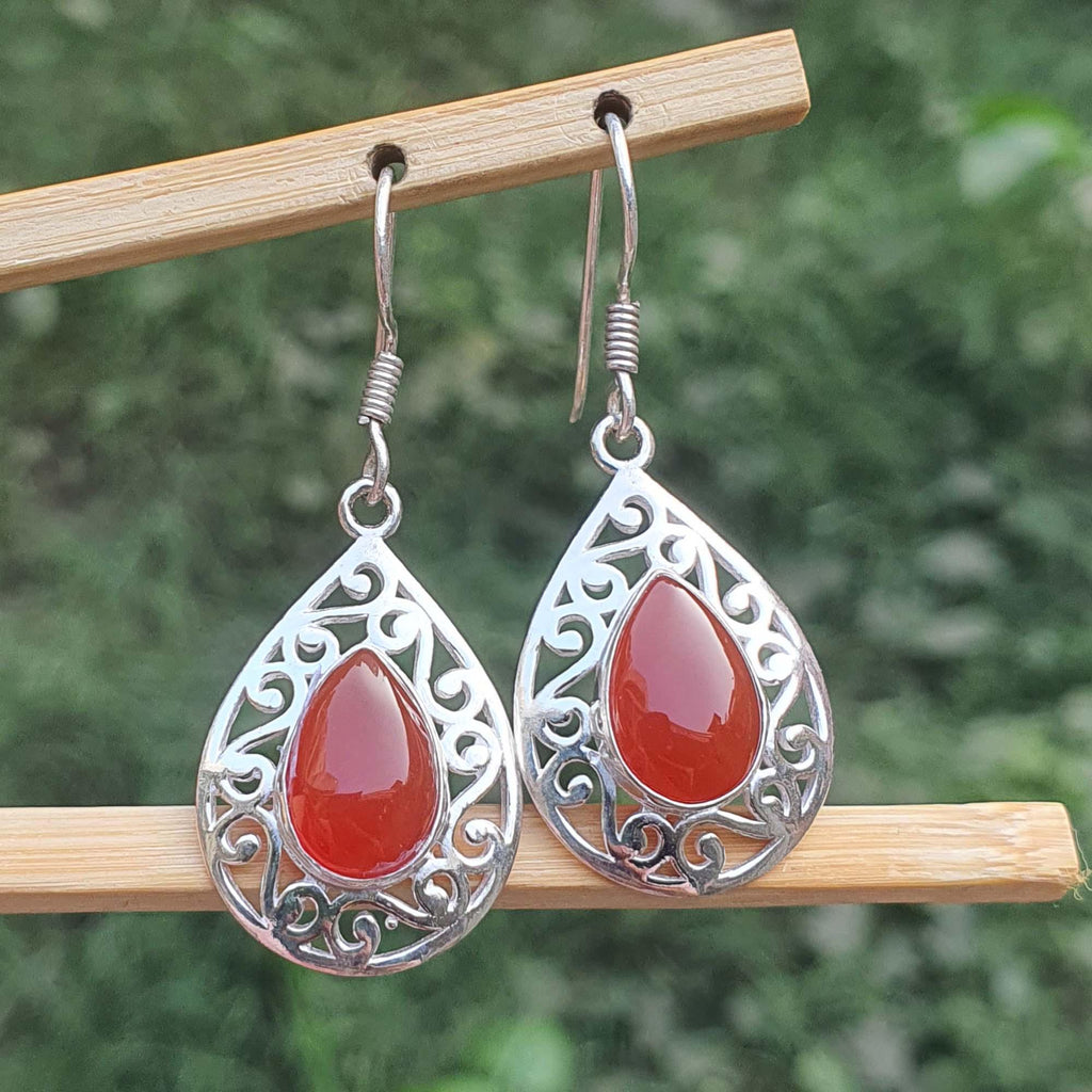 Teardrop-shaped earrings with red stones and silver filigree on a wooden stick against a blurred green background.