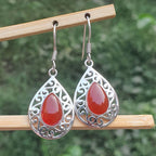 Teardrop-shaped earrings with red stones and silver filigree on a wooden stick against a blurred green background.