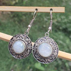Pair of silver earrings with circular designs and light-colored stones on a wooden stick against a blurred green background.