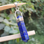 Blue lapis lazuli pendant with silver bail on a wooden stick against a green leafy background