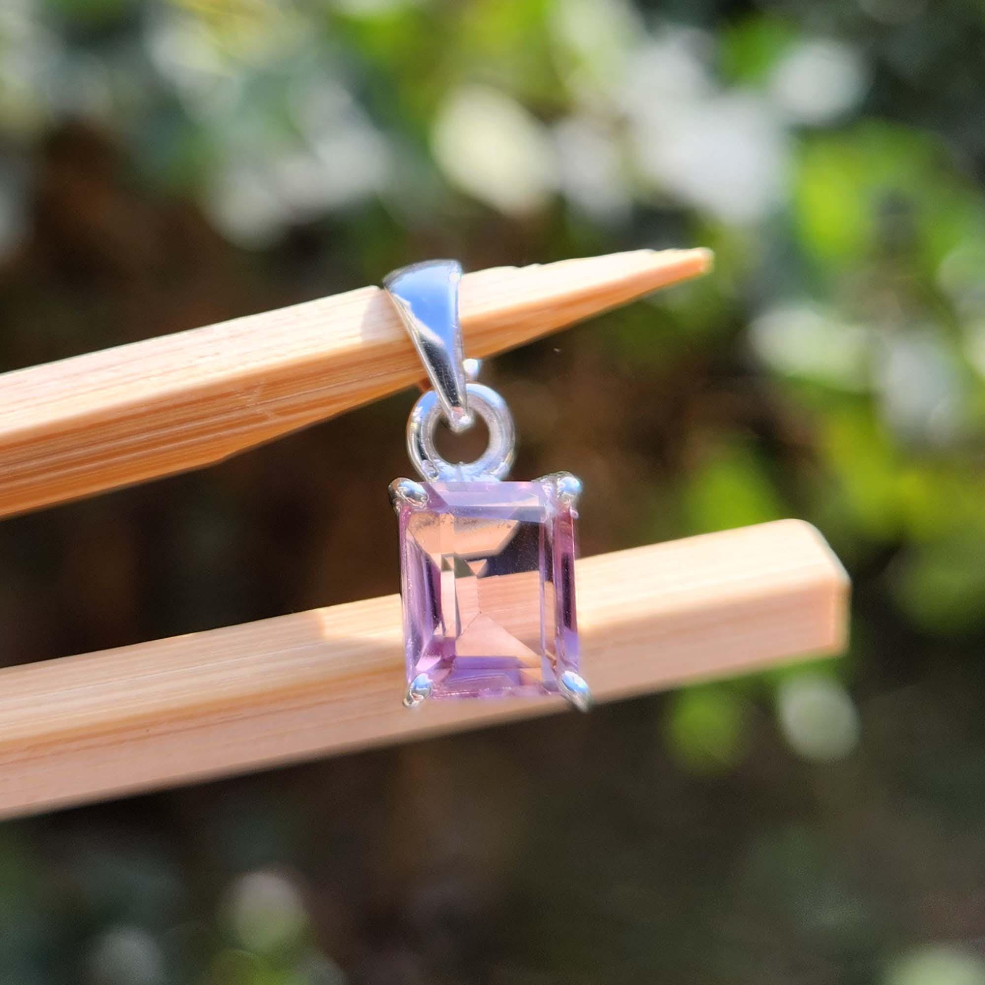 Silver pendant with a pink gemstone held by wooden tweezers against a blurred green background