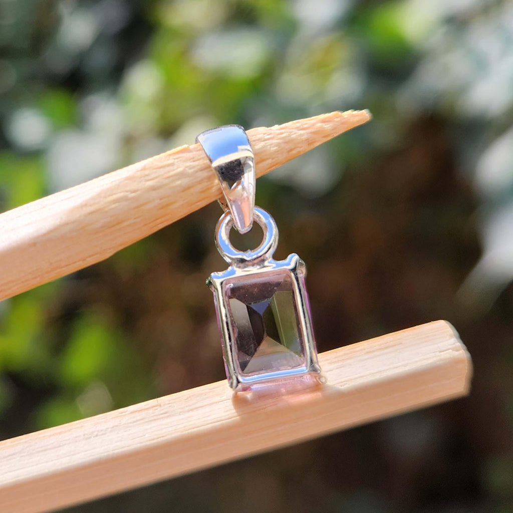Silver pendant with a rectangular stone on a wooden stick against a blurred natural background