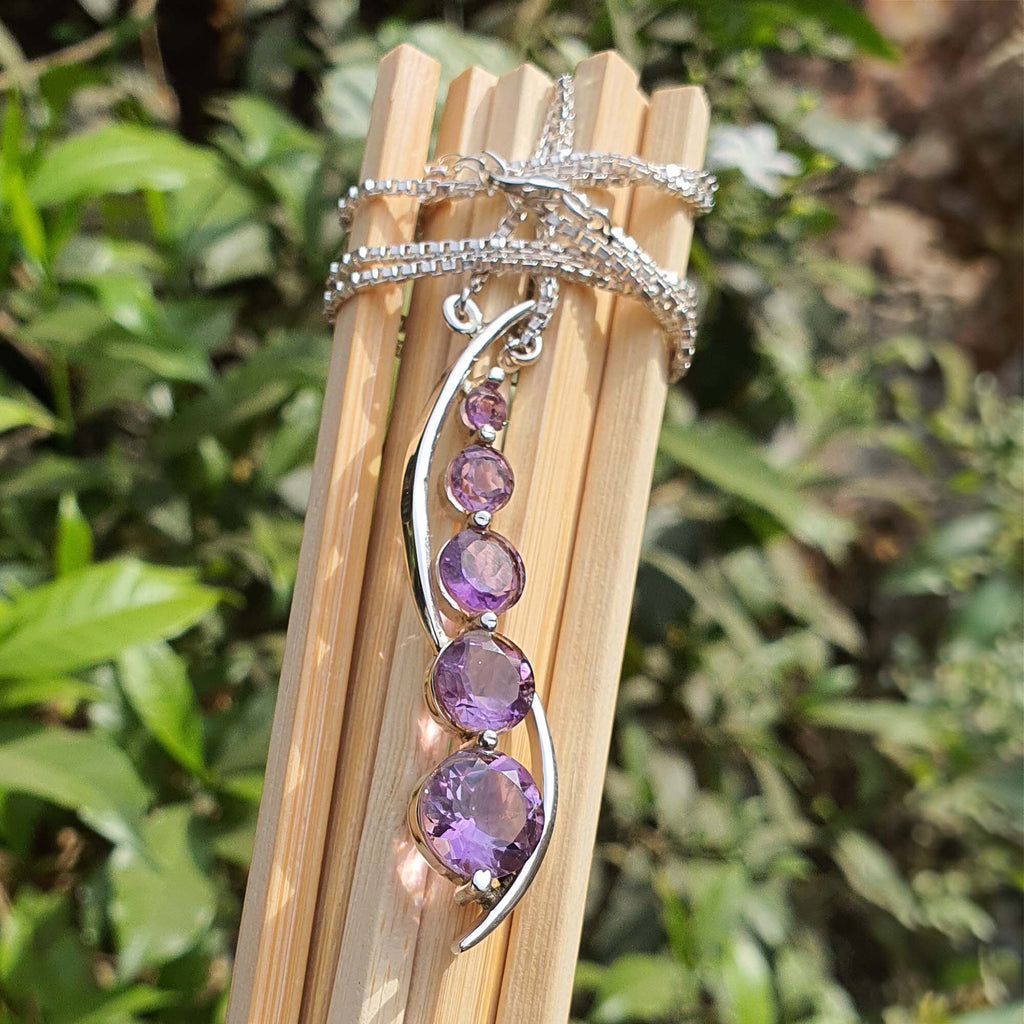 Silver necklace with purple gemstones on a wooden stand against a green leafy background
