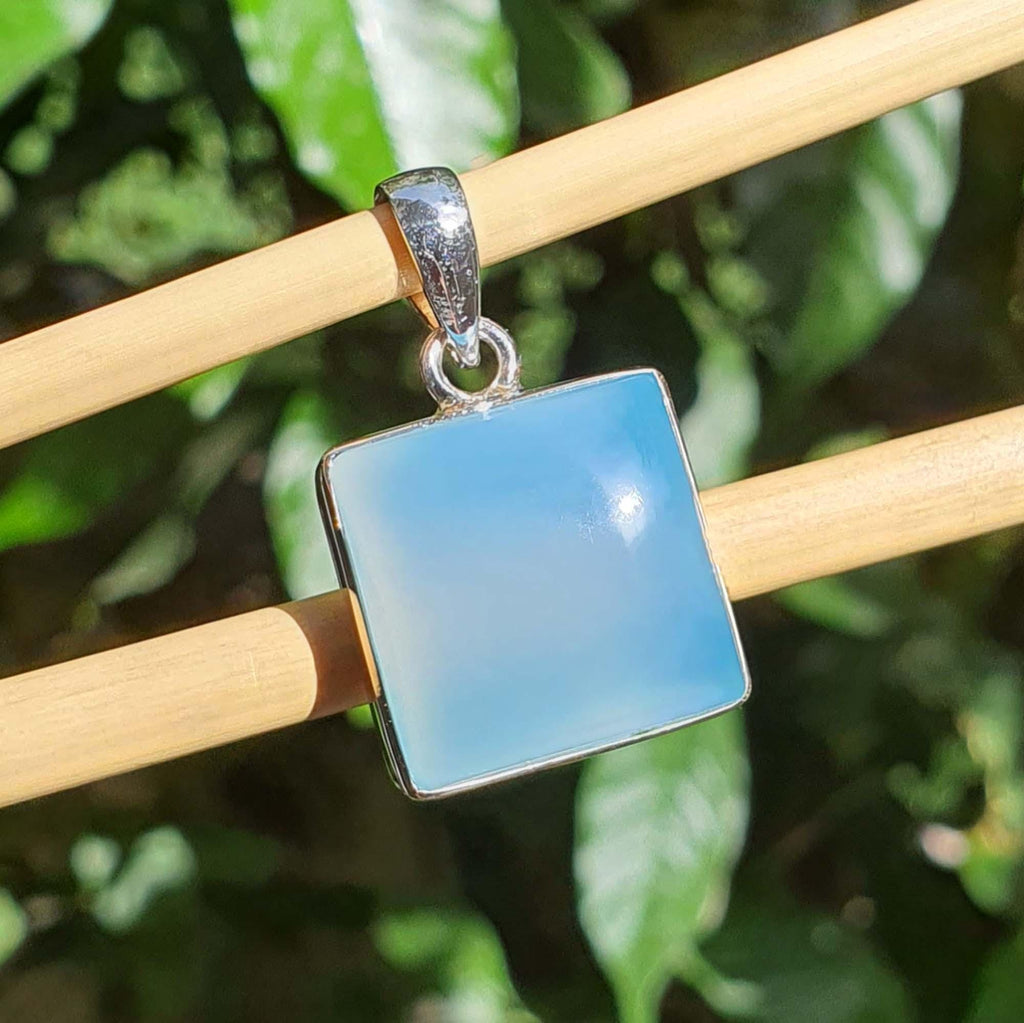 Blue square pendant with silver frame on a wooden stick against a green leafy background