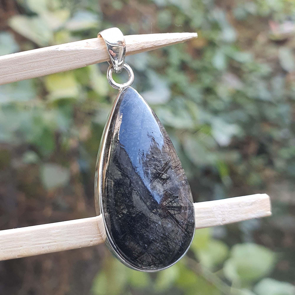 Teardrop-shaped pendant with a dark stone and silver frame on a stick against a blurred green background
