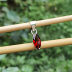 Silver pendant with a red gemstone on a wooden stick against a green foliage background