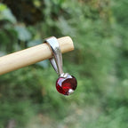 Silver pendant with a red gemstone held by a wooden stick against a blurred green background